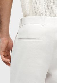 Άνδρας που φοράει λευκό παντελόνι με ραφή στο πίσω μέρος και θηλιές για ζώνη, συνδυασμένο με λευκό υφασμάτινο αμάνικο τοπ.