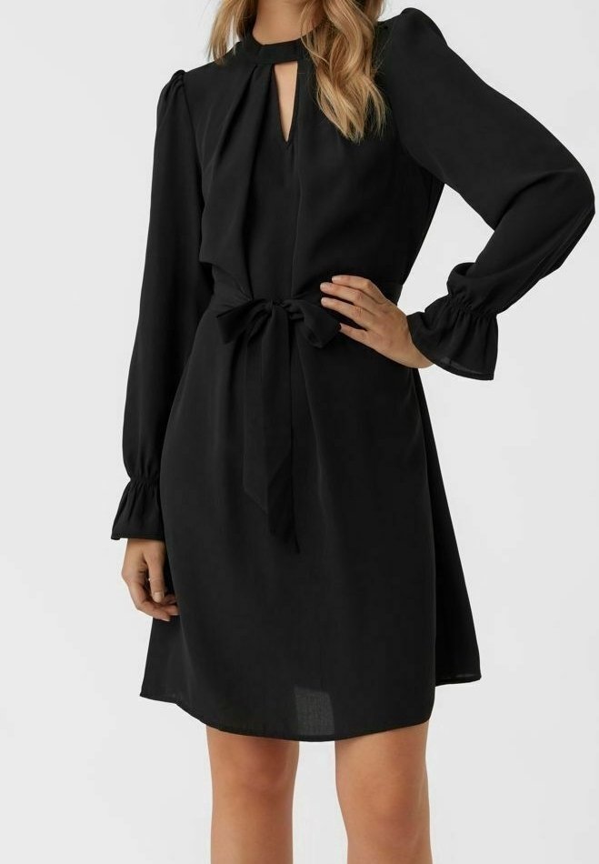 Femme portant une robe noire à manches longues avec un décolleté en forme de goutte, des poignets à volants et une ceinture nouée à la taille, debout avec une main sur la hanche.