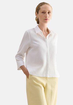 Frau mit heller Haut und zurückgebundenen Haaren, trägt eine weiße Bluse mit langen Ärmeln und hellgelbe Hose, steht mit den Händen in den Taschen.