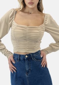 Beige gerafftes Langarmshirt mit eckigem Ausschnitt. Der Stoff ist glatt und eng anliegend, kombiniert mit hoch taillierten Jeans.