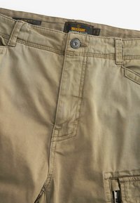 Uske slim fit kahedi cargo hlače s preklopnim zatvaranjem, ojačanom šavovima i prednjim zakrpama. Tkanina je glatka s laganim sjajem.