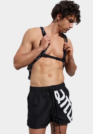 Mężczyzna bez koszulki regulujący paski plecaka z czarnym logo, ubrany w czarne kąpielówki z dużymi białymi literami na lewej nodze.