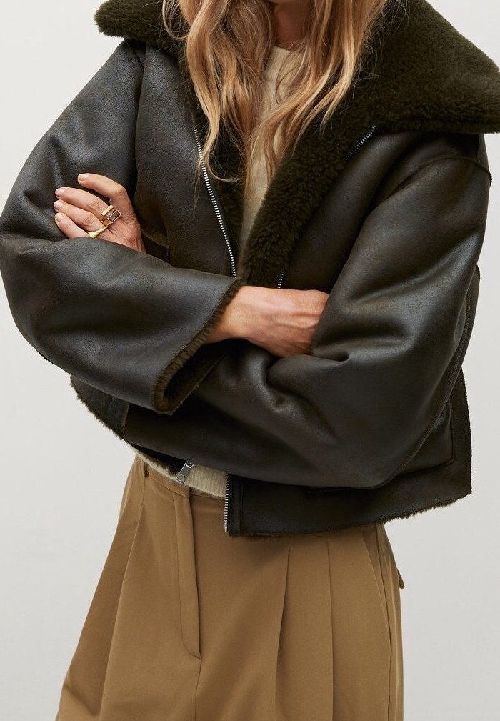 Femme portant une veste en peau lainée noire avec un grand col par-dessus un pull beige et un pantalon marron taille haute, bras croisés.