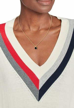 Femme portant un pull blanc à col en V avec des rayures rouges, grises et bleu marine et un collier en or avec un pendentif circulaire noir.