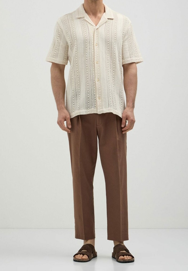 Homme portant une chemise en maille crème à manches courtes et boutons, un pantalon marron court et des sandales marron foncé, debout devant un fond clair uni.