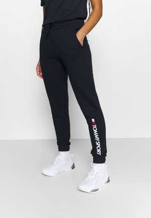 Personne portant un jogging noir Tommy Sport avec un logo sur le bas de la jambe et des baskets blanches, debout devant un fond clair.