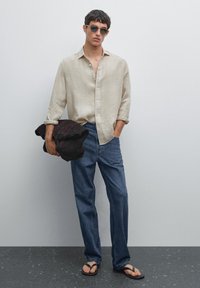 Man wearing light beige buttoned shirt, blue jeans, black flip-flops, sunglasses, holding dark brown folded jacket against plain wall.