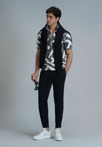 Grau gemusterte Polo mit kurzen Ärmeln und schwarzen Akzenten, marineblauer ärmelloser Pullover, marineblaue tapered Hose und weiße Sneakers; hält schwarze Sonnenbrillen.