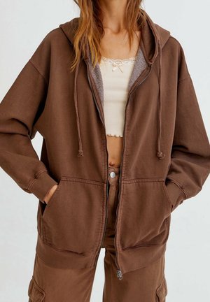 Žena nosící hnědou mikinu na zip s rukama v kapse, přes bílý krajkový crop top a odpovídající hnědé kalhoty.