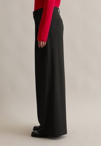 Pantalones negros de pierna ancha con acabado texturizado, que presentan una cintura alta y bolsillos laterales, combinados con un suéter ajustado de color rojo brillante.