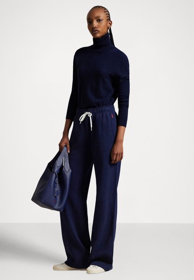 Granatowy sweter z golfem, luźne granatowe spodnie z białym sznurkiem i granatowa torebka z fakturą. Model nosi białe trampki.