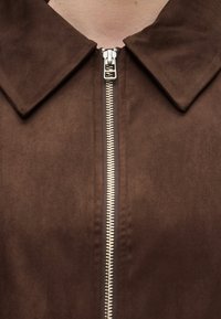 Κοντινό πλάνο καφέ σαλονιού σακακιού με ασημένιο μεταλλικό φερμουάρ που τρέχει κάθετα κάτω από γιακά με αιχμηρό σχήμα.