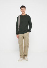 Junger Mann steht vor weißem Hintergrund und trägt einen dunkelgrünen Strickpullover, beige Cargo-Hosen, weiße Sneakers und eine Umhängetasche.