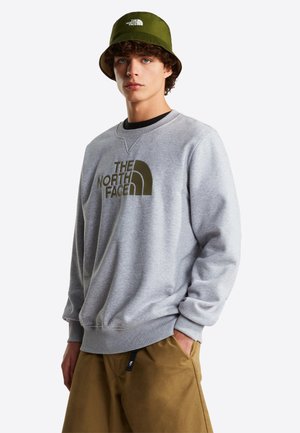 Junger Mann, der einen olivgrünen Bucket Hat und ein graues Sweatshirt mit The North Face-Logo trägt, kombiniert mit braunen Hosen, steht mit einer Hand in der Tasche.