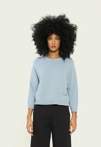Pulover oversized de culoare albastru deschis, cu guler rotund, mâneci 3/4 și o mică buzunar frontal. Textură moale și detalii cu tiv striat.