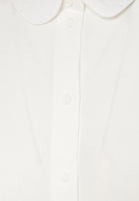 Närbild av vit texturerad tyg med tre vita knappar och en rundad krage på ett plagg.