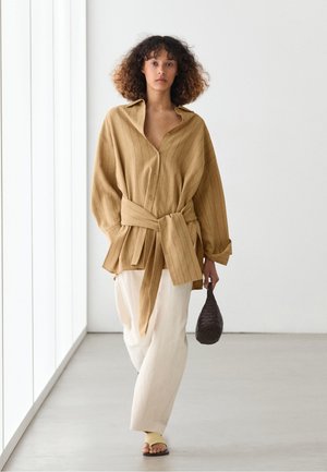 Vrouw met krullend haar die een losse beige blouse met knopen en een gestrikte taille draagt, crème brede broek, sandalen, en een kleine donkere geweven tas vasthoudt.