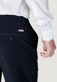 Pantaloni navy in tessuto morbido, con chiusura a bottoni e una piccola etichetta metallica del marchio sulla vita. Camicia bianca con polsini a bottone.