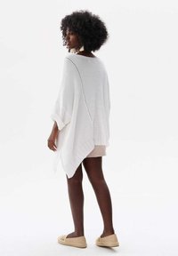 Weißes Strickponcho mit weiten Ärmeln, strukturiertem Design und V-Ausschnitt. Über einem beigen Kleid getragen, kombiniert mit hellbraunen Espadrilles.