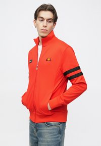 Punane tõmblukuga jakk, valmistatud siledast kangast, millel on kõrge krae, varrukatel musta triibuga aktsendid ja rinnal logomärgis.