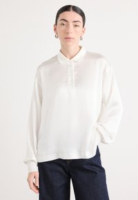 Chemise polo blanche à manches longues en tissu lisse, dotée d'un col à boutons et d'un subtil détail brodé sur la poitrine.