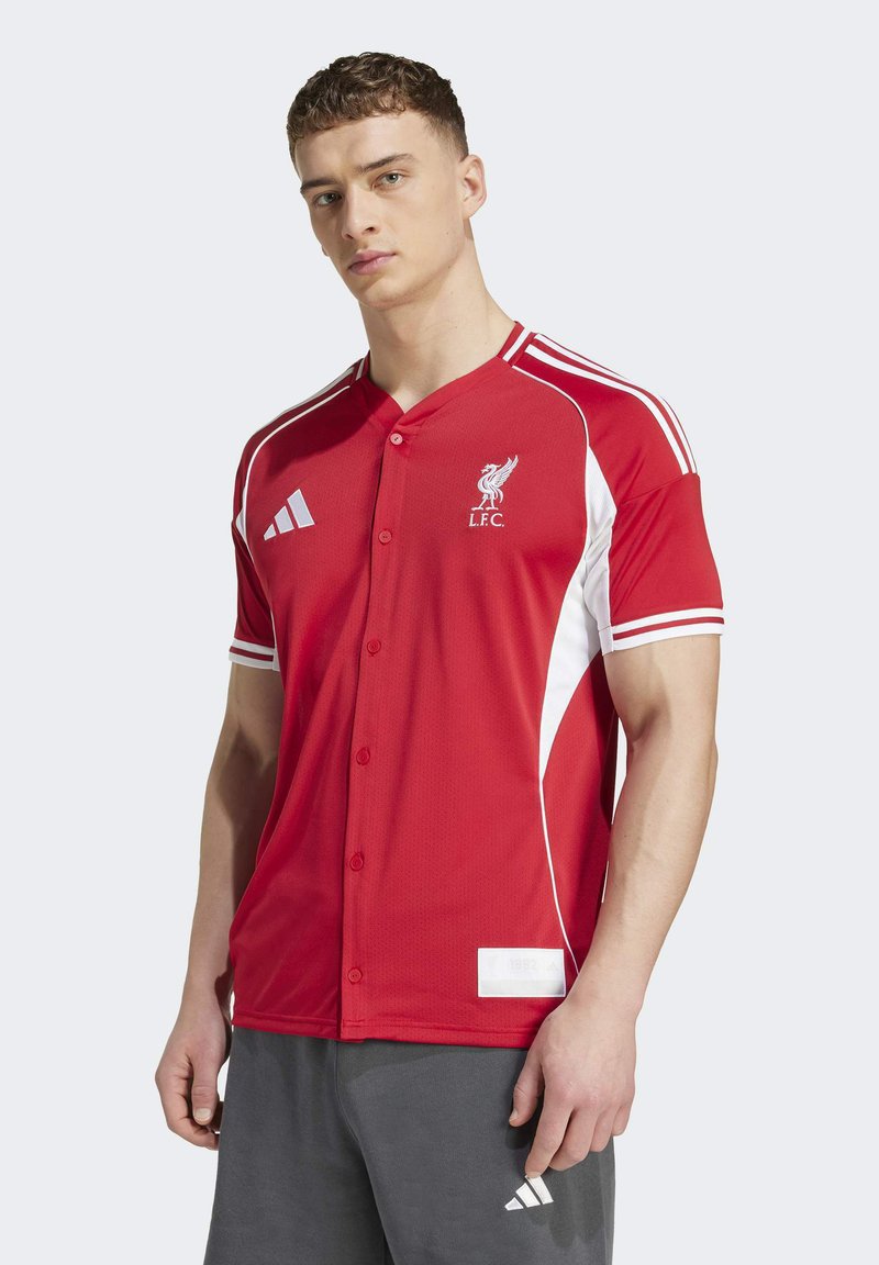 Ung man i en röd Liverpool FC-sporttröja med vita ränder och grå Adidas-shorts mot en enfärgad bakgrund.