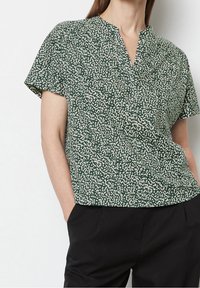 Blusa verde a maniche corte con scollo a V, caratterizzata da un motivo bianco maculato. Tessuto leggero, vestibilità morbida, indossata con pantaloni neri.