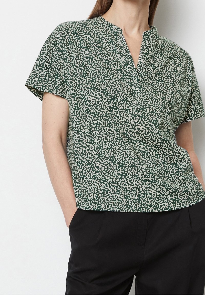 Blusa verde a maniche corte con scollo a V, caratterizzata da un motivo bianco maculato. Tessuto leggero, vestibilità morbida, indossata con pantaloni neri.