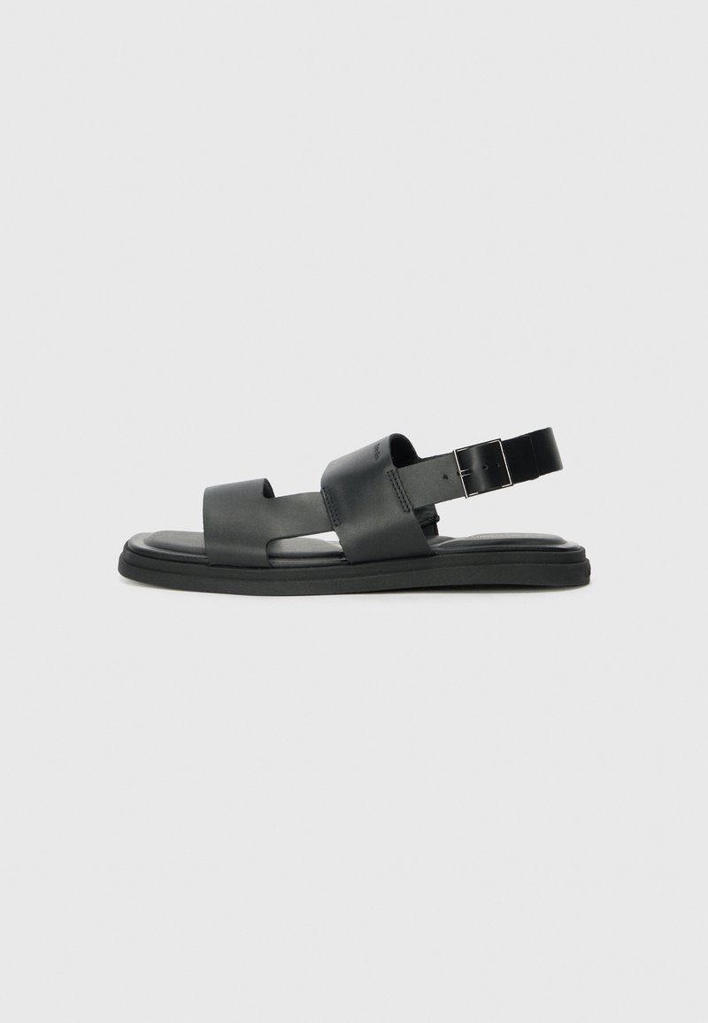 Black leather sandals with a flat sole, featuring two wide straps across the foot and an adjustable ankle strap with a metal buckle.