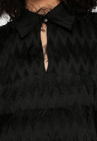 Μαύρο μπλουζάκι με υφή και ζιγκ-ζαγκ μοτίβα, μικρή λαιμόκοψη σε σχήμα κλειδαρότρυπας με κουμπί και μεγάλο γιακά.