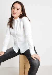 Weißes Langarm-Hemd mit Knopfleiste und figurbetontem Design, kombiniert mit dunklen Jeans. Glatte Textur, klassischer Kragen und seitlicher Splitleisten-Detail.