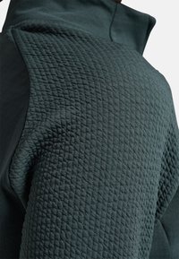 Mørkegrøn træningsjakke med tekstureret, quiltet stof på skulderen og glat materiale på overarmen. Høj krave design.