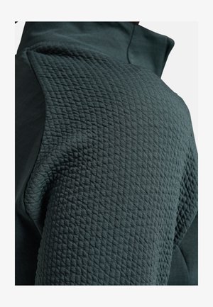 Dunkelgrüne Sportjacke mit strukturiertem, gestepptem Stoff auf der Schulter und glattem Material am Oberarm. Hohe Kragenform.