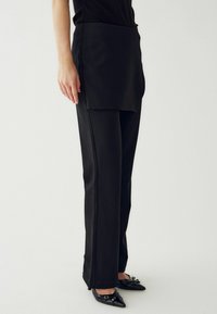 Melnas platās bikses ar gludu audumu, kurām ir priekšējais pārklājums un sānu šuves. Saskaņotas ar melnām kurpēm ar smailu purngalu.