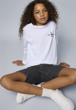 T-shirt blanc à manches longues avec un petit logo noir, associé à une jupe en denim grise et des baskets blanches avec des accents texturés.