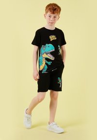 Garçon aux cheveux roux portant un ensemble short et t-shirt noir avec un dinosaure T-Rex coloré et le texte "SUPER SKATER", debout sur un fond jaune.