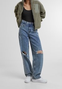 Flauschige olivgrüne Bomberjacke mit Reißverschlusstasche, kombiniert mit hellblauen Jeans mit Rissen und weißen Sneakern.