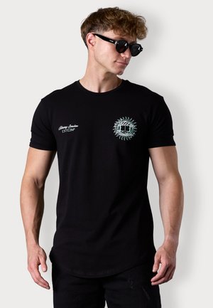 T-shirt en coton noir avec un design graphique sur la poitrine gauche, encolure ronde et manches courtes. Porté avec des lunettes de soleil noires.