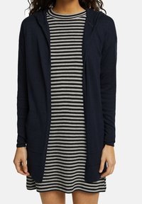 Marine gebreide cardigan met capuchon, open voorkant en twee voorzakken, gelaagd over een gestreepte zwart-witte jurk met korte mouwen.