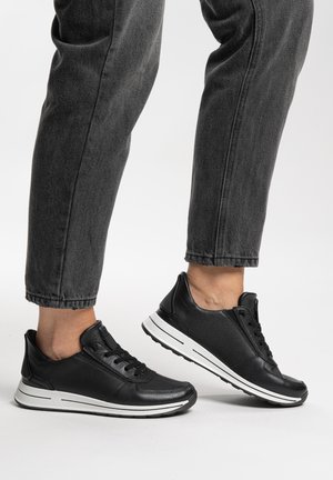 Schwarze Ledersneaker mit weißen Sohlen, getragen zu dunkelgrauen, hochgekrempelten Jeans, vor weißem Hintergrund.