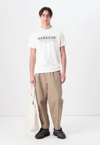 Λευκό βαμβακερό T-shirt με μαύρο κείμενο, μπεζ φαρδιά παντελόνια, μαύρα παπούτσια και μια κρεμ τσάντα ώμου. Απλό φόντο.