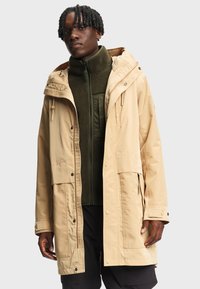 Homme aux cheveux noirs tressés portant un imperméable à capuche beige par-dessus une veste en polaire vert olive sur un fond uni clair.
