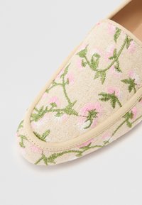 Beige színű, hímzett slip-on cipő rózsaszín virágmintákkal és z�öld levelekkel, strukturált, puha felülettel és semleges szegélyezéssel.