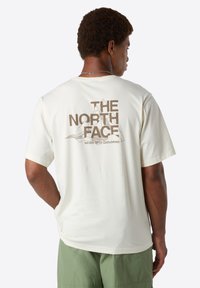 Homme portant un t-shirt crème avec "THE NORTH FACE" et un graphique de montagne au dos, associé à un short vert, tourné dos à l'appareil photo.
