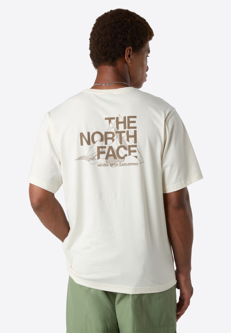 Homme portant un t-shirt crème avec "THE NORTH FACE" et un graphique de montagne au dos, associé à un short vert, tourné dos à l'appareil photo.