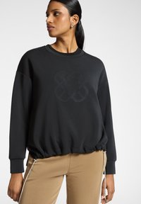 Schwarzer Pullover mit einer strukturierten, geprägten Gestaltung in der Mitte und gerafftem Saum. Lange Ärmel und weicher Stoff. Kombiniert mit beigen Hosen.