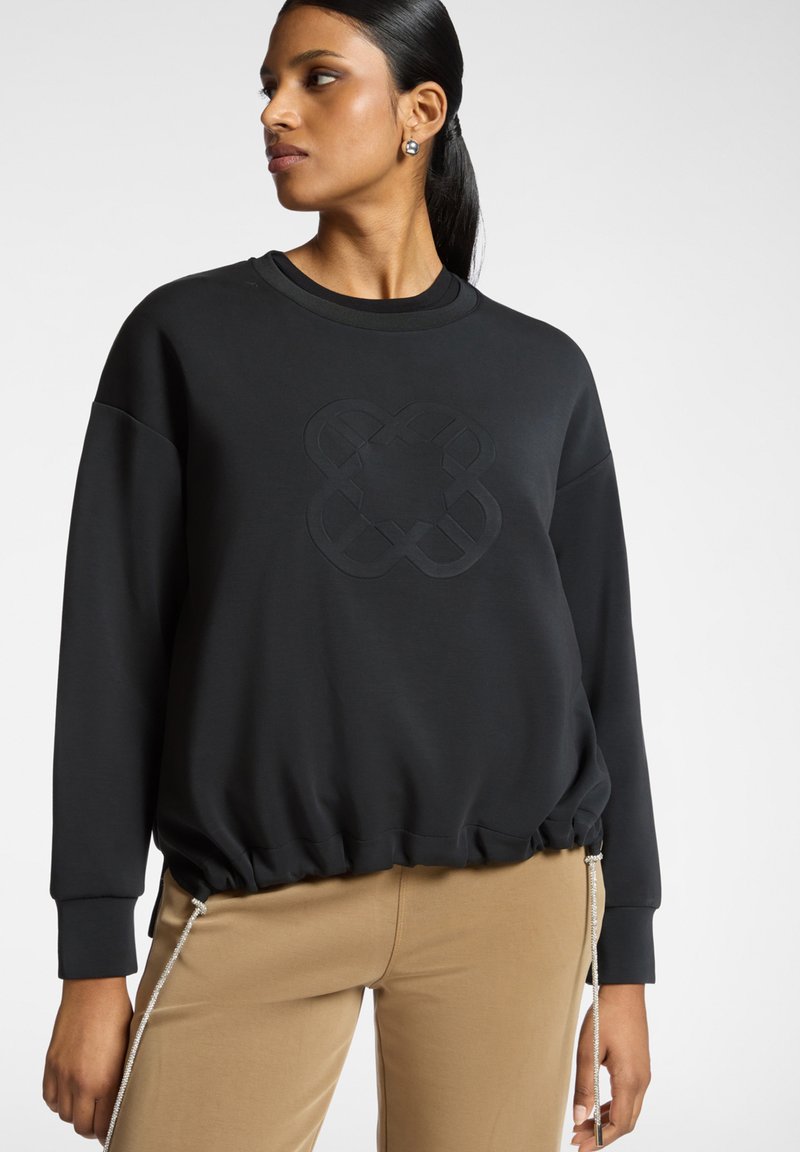 Schwarzer Pullover mit einer strukturierten, geprägten Gestaltung in der Mitte und gerafftem Saum. Lange Ärmel und weicher Stoff. Kombiniert mit beigen Hosen.