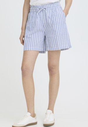 Ember fehér és kék függőleges csíkos, megkötős derekú rövidnadrágban és fehér tornacipőben, sima háttér előtt áll.