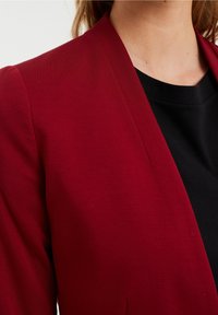 Blazer rojo con un diseño suave y estructurado, con cuello chal, combinado con una camiseta negra de cuello redondo debajo.