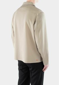 Beige overhemd met lange mouwen en een kraag, gladde textuur en subtiele oranje branding. Aansluitend ontwerp, van de achterkant weergegeven met zwarte broek.